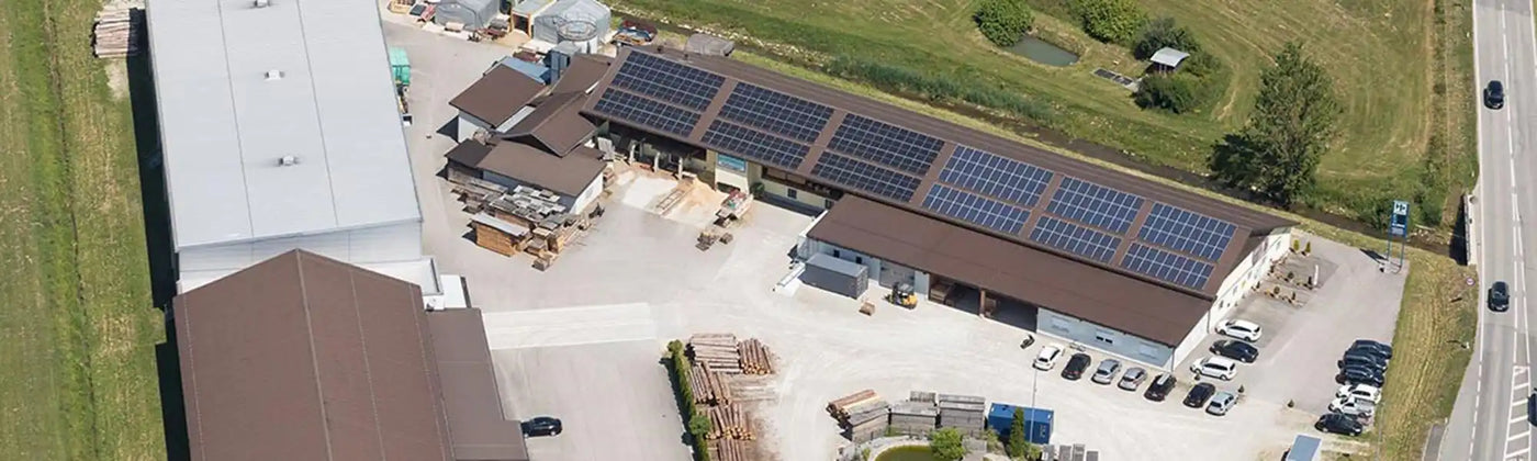 Luftaufnahme einer Gewerbeimmobilie mit mehreren Gebäuden, Solarpanelen, einem großen Parkplatz und einem Außenlagerbereich für verschiedene Materialien.