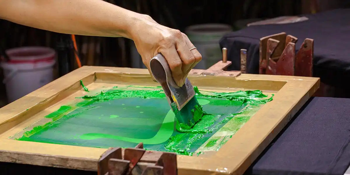 Die Hand einer Person, die einen Rakel benutzt, um grüne Tinte auf eine Stoffoberfläche zu screenprinten.