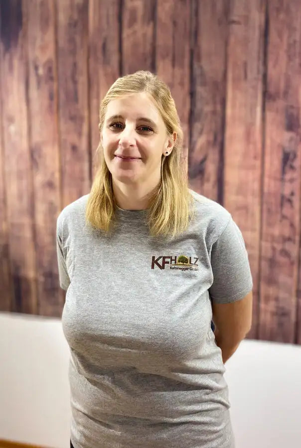 Graues, heathered Baumwoll-T-Shirt mit einem kleinen gestickten Logo auf der linken Brust, das ’kfholtz’ in schwarz-grüner Schrift über einer stilisierten Baumgrafik zeigt.