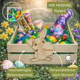 Ein hellfärbiger hölzerner Ostkorb mit geschnitzter Bunny-Dekoration, die den Namen »Verena« hält, gefüllt mit bunten Schokoladeneiern, Riegeln und grünem Konfetti.