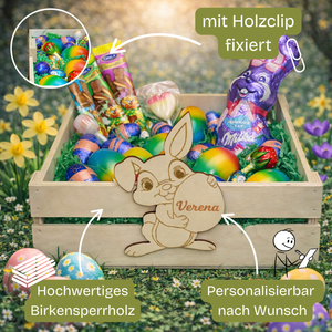 Ein hellfärbiger hölzerner Ostkorb mit geschnitzter Bunny-Dekoration, die den Namen »Verena« hält, gefüllt mit bunten Schokoladeneiern, Riegeln und grünem Konfetti.