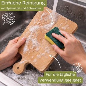 Hellbraunes hölzernes Schneidebrett mit Griff und abgerundeten Ecken, das unter fließendem Wasser mit einem grün-gelben Schwamm geschrubbt wird.